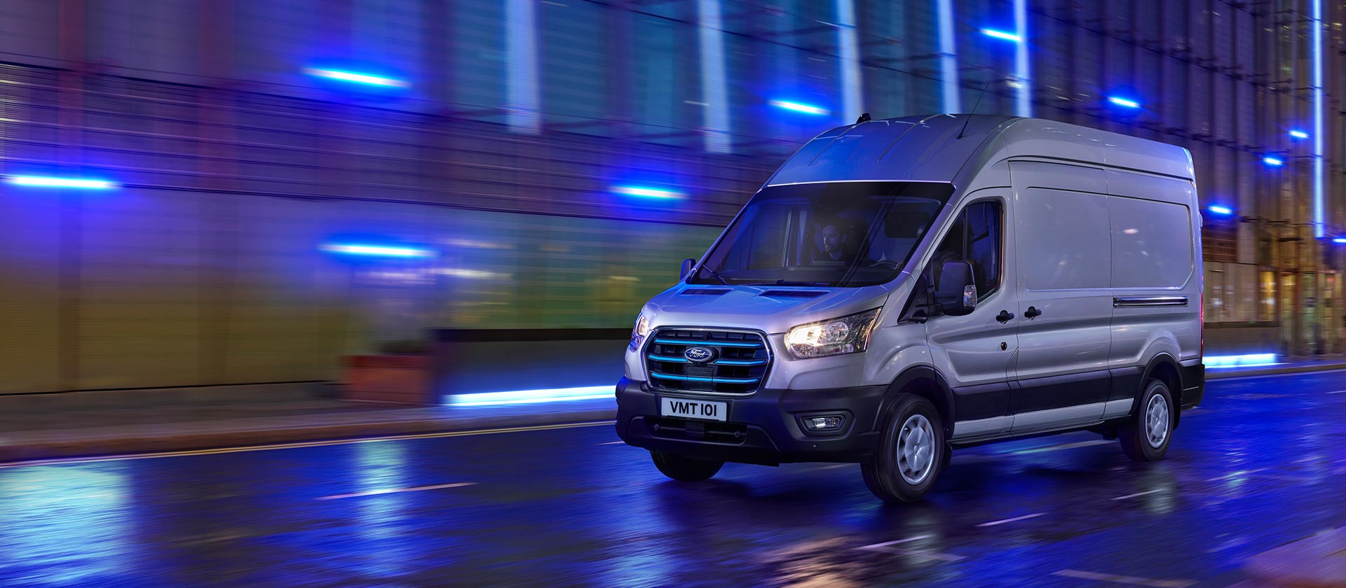 Silver Ford E-Transit electric van driving on a wet city street at night.
