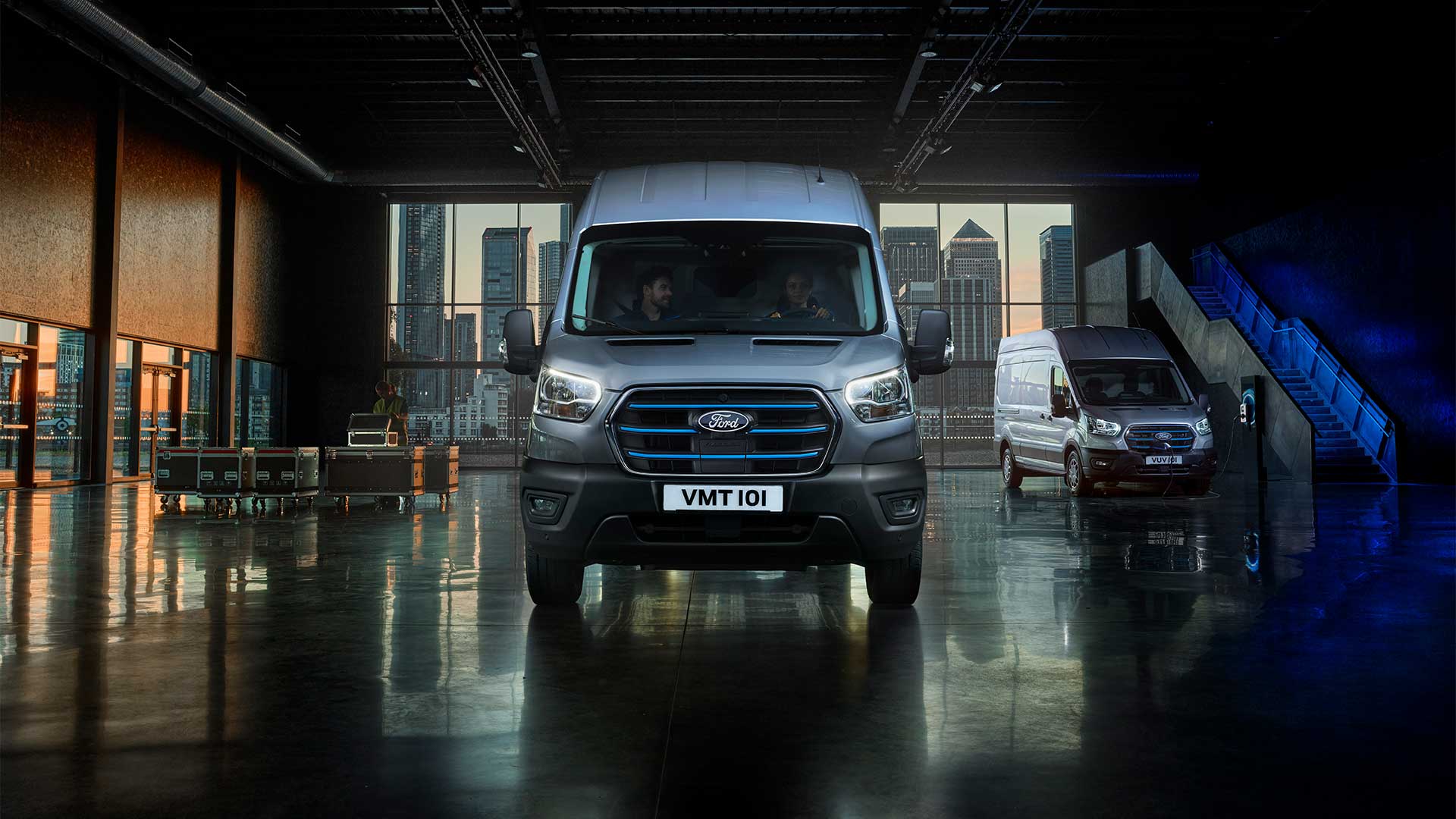 Front view of two Ford Transit vans parked in a garage with a city skyline through the windows.