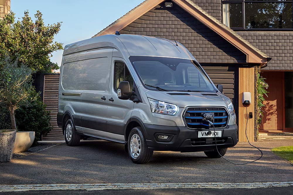 Silver Ford E-Transit van parked in a driveway next to a house, connected to a charging station.