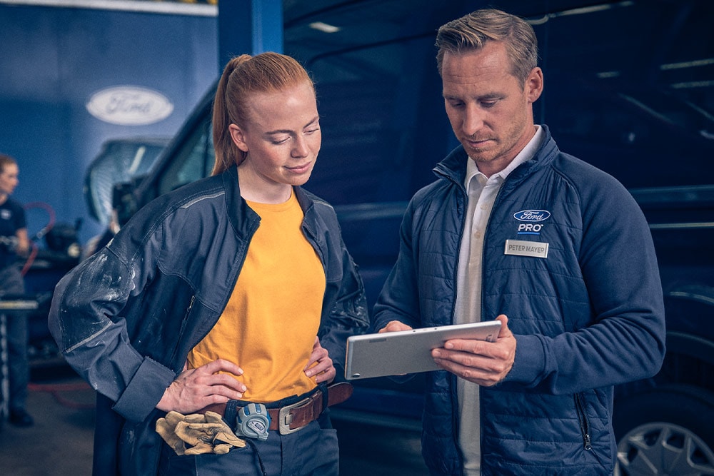 Two mechanics in a garage looking at a tablet in front of a Ford E-Transit electric van.