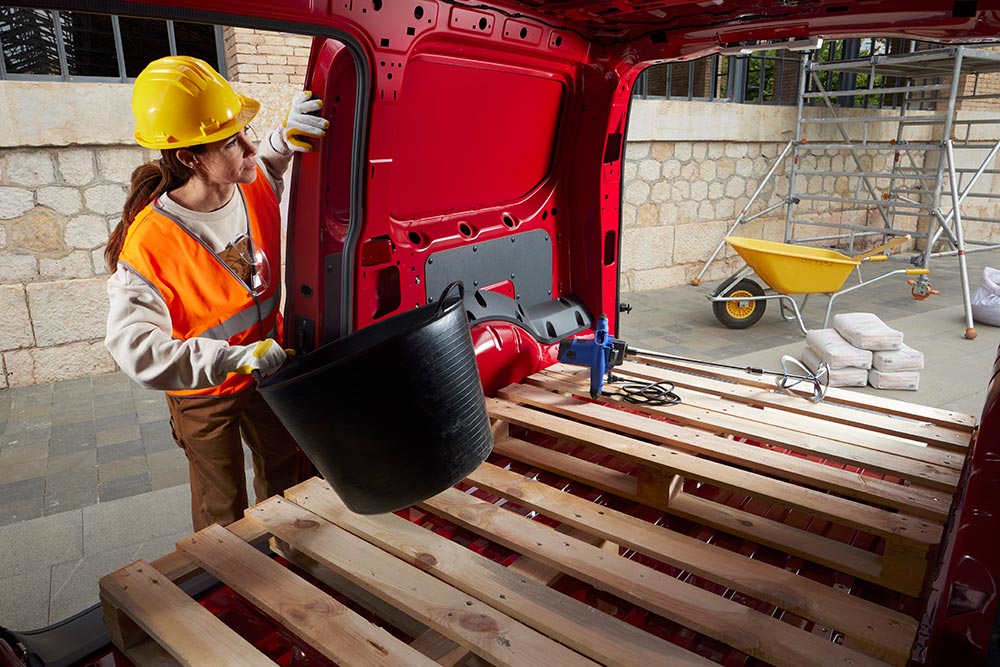 A woman puts construction materials in the loadspace of a Ford Transit Connect, with side and rear doors open.