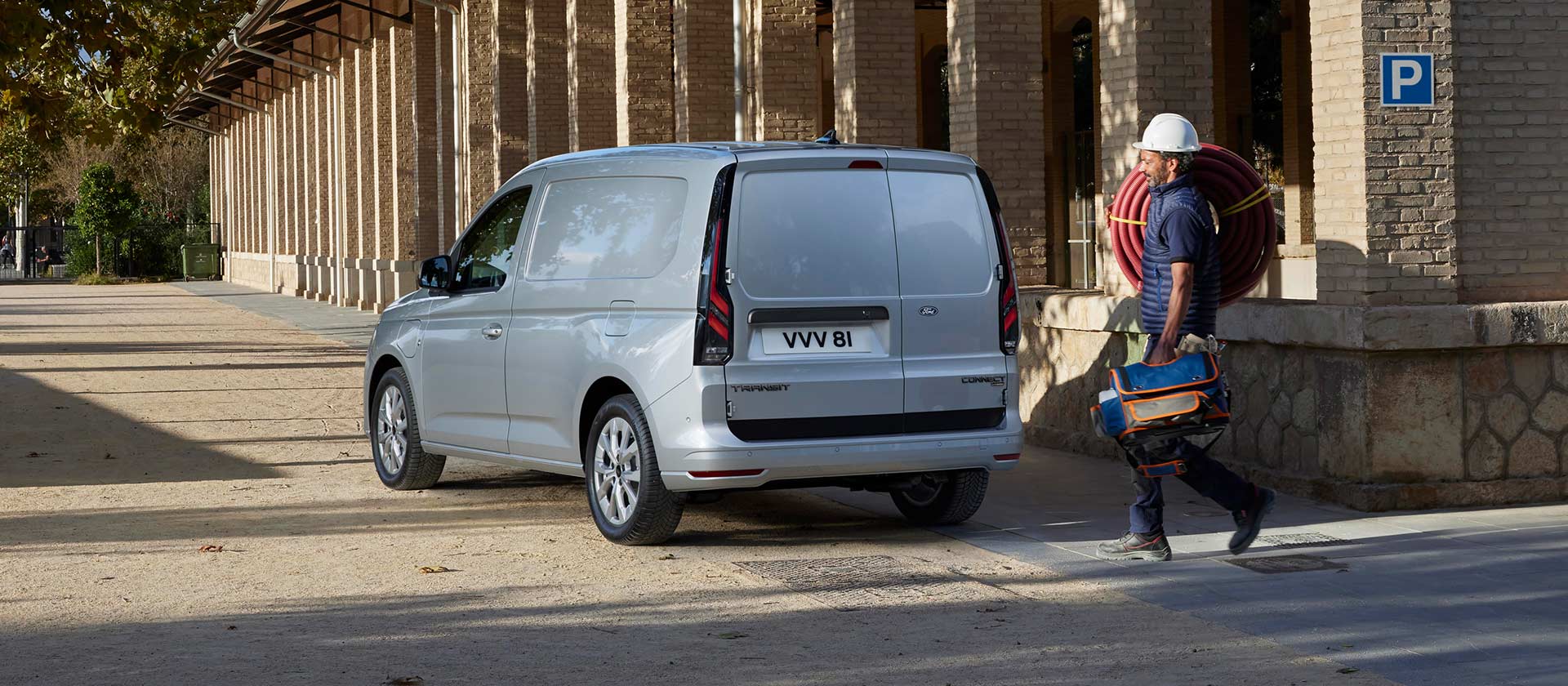 A worker walking next to a parked silver Ford Transit Connect.