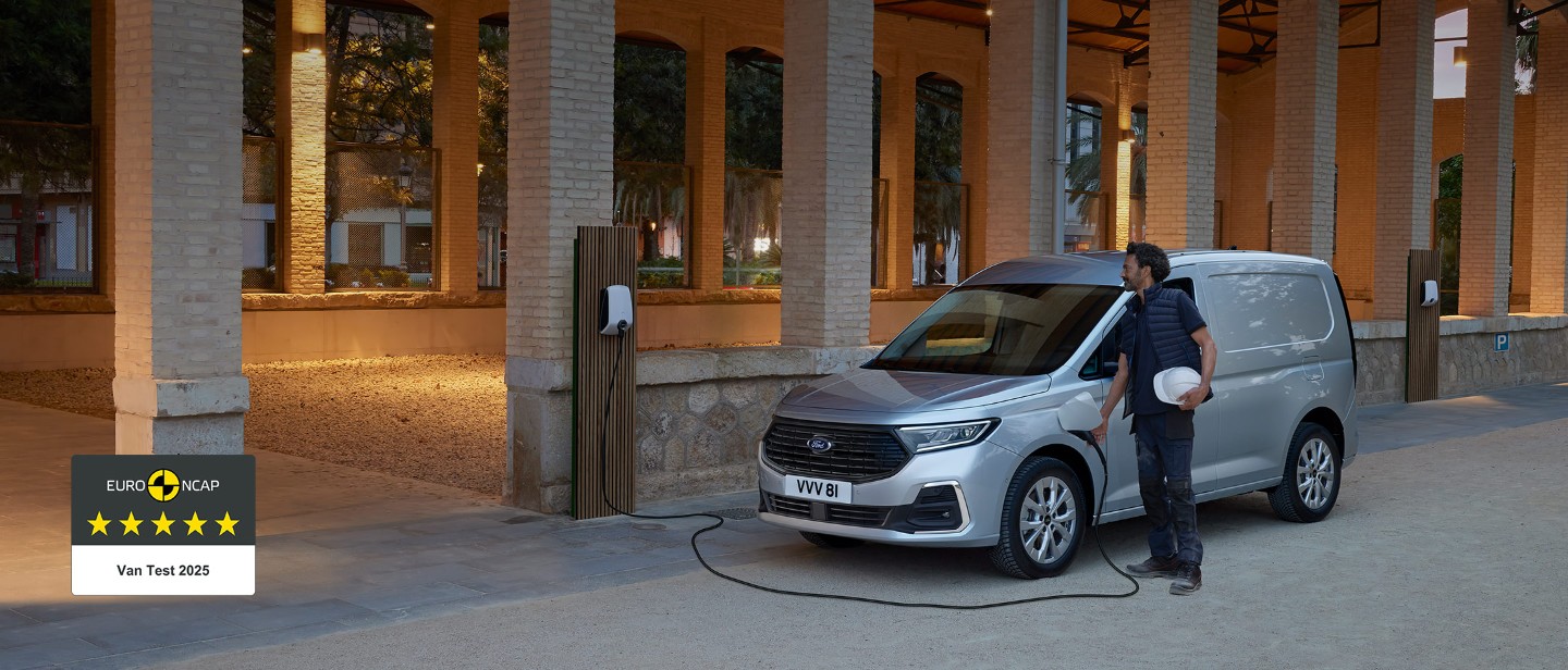 A man holding a hard hat plugs a wall box charging cable into Ford Transit Connect in Stardust Silver.
