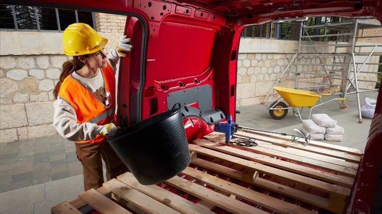 A woman places materials into the loadspace of Ford Transit Connect in Maple Red with the side and back doors open, on a building site.