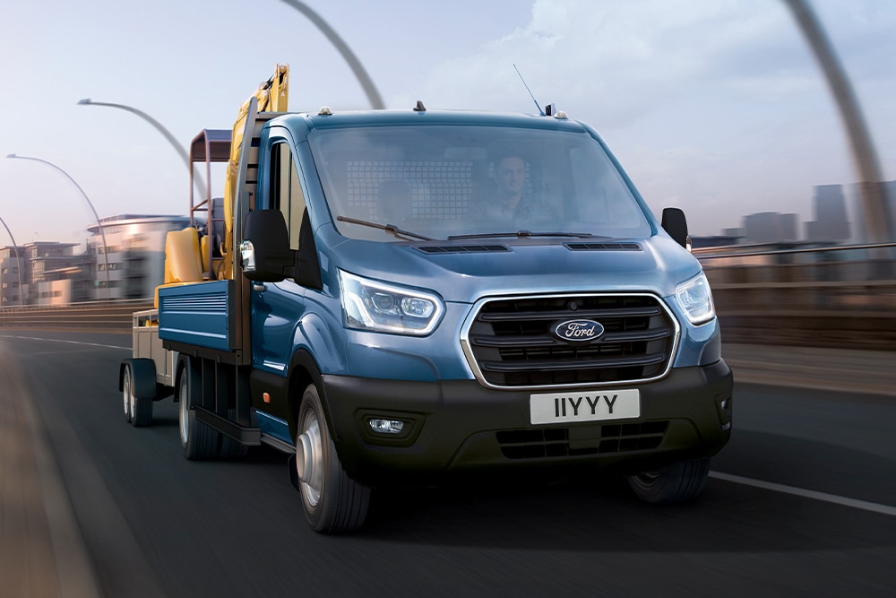  Front grille view of a Ford Transit Chassis Cab carrying a digger while driving along a city road.