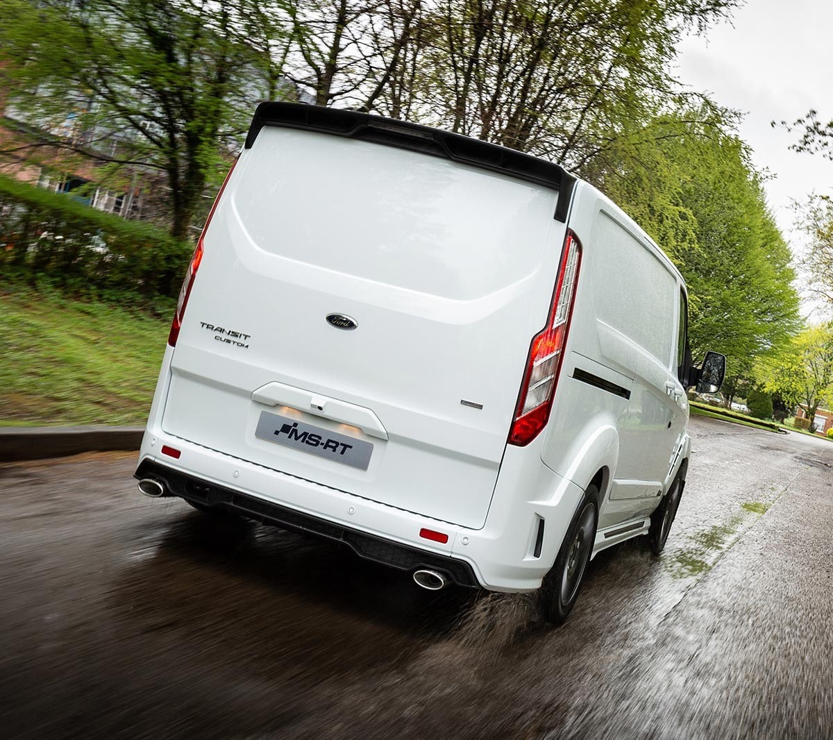 White Ford Transit Custom MS-RT driving down a road next to a wooded area