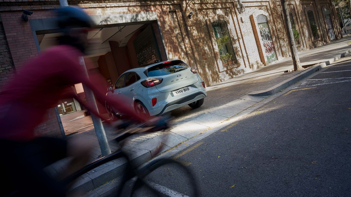 A grey Ford Puma reversing out of a driveway while a cyclist wearing red passes by.