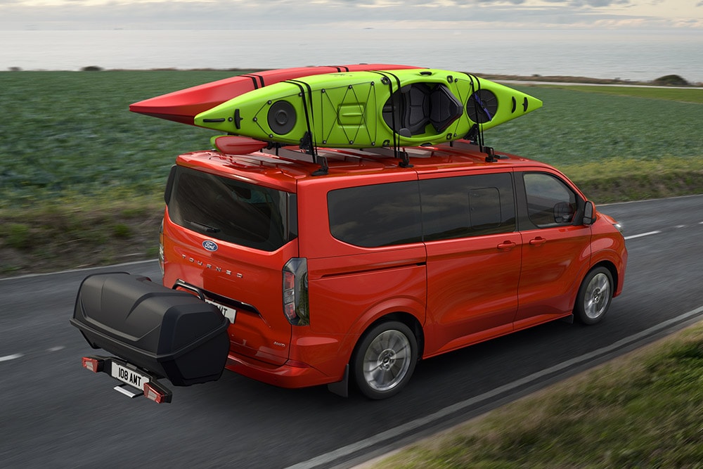 Red Ford Pro Transit with two kayaks on the roof, towing a storage carrier, and driving along a coastal road.
