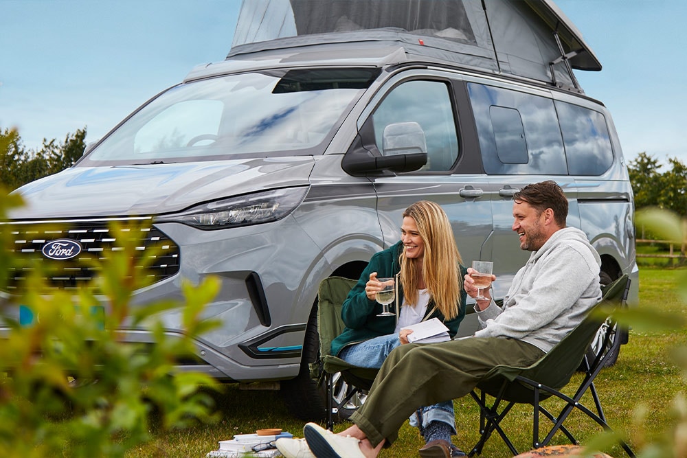 A couple enjoying drinks in camping chairs next to a grey Ford Transit Custom camper van with a pop-up roof.