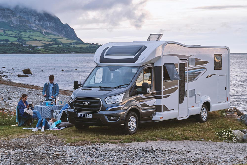 A grey and white Ford Transit motorhome parked by a lake, with a family having a picnic.