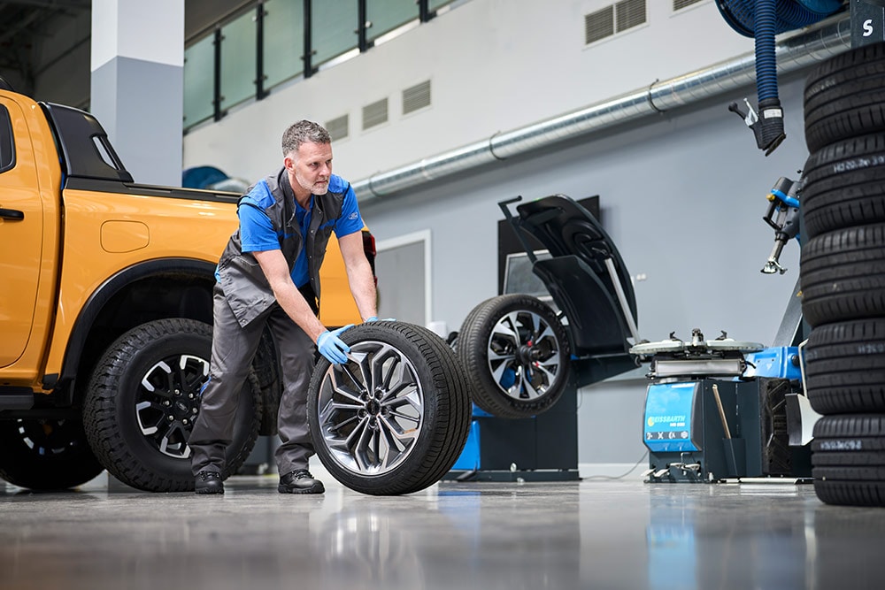 A Ford mechanic wheeling a tyre across a warehouse, a yellow Ford truck parked in the background.