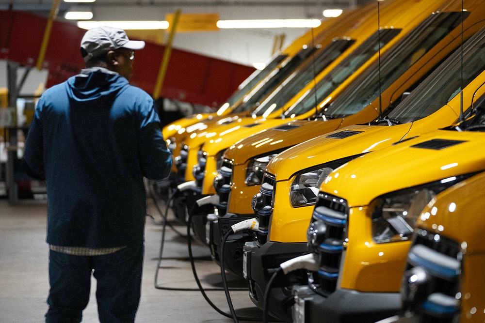 A row of yellow Ford electric vans being charged, a man is standing next to them.