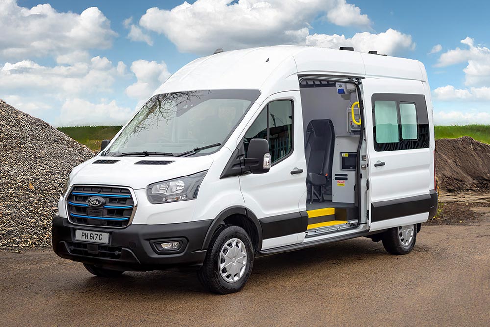 A white Ford E-Transit van with its side door open, parked outdoors near gravel piles under a cloudy sky.