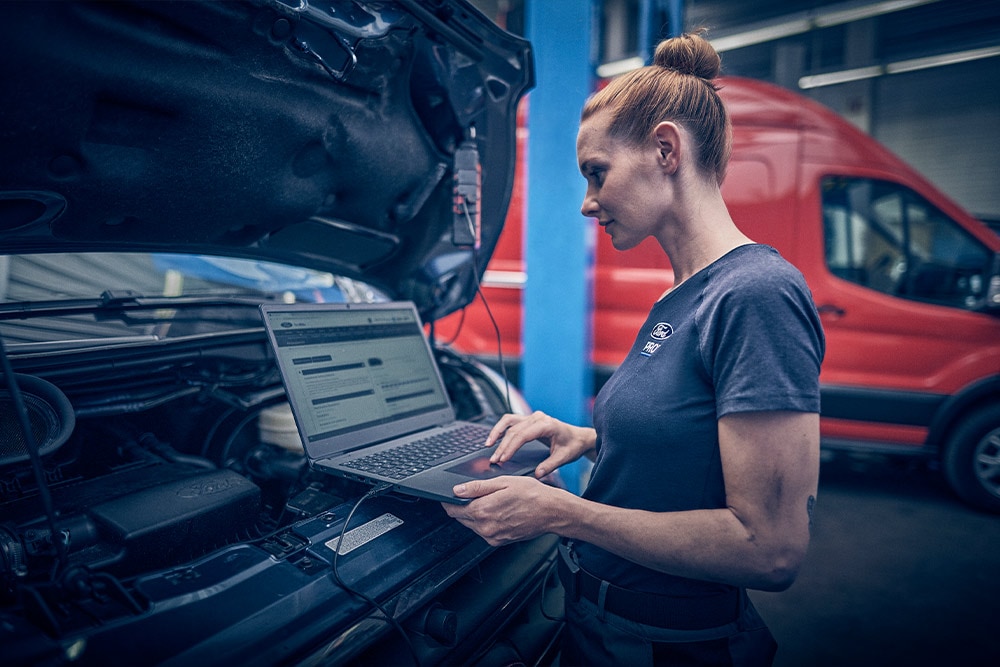 A woman using a laptop connected to the engine bay of a Ford van in a garage.