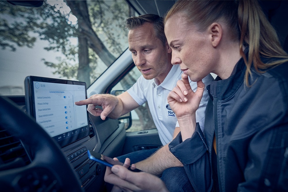A man pointing to a large touchscreen inside a van, while a woman looks on, holding a smartphone.