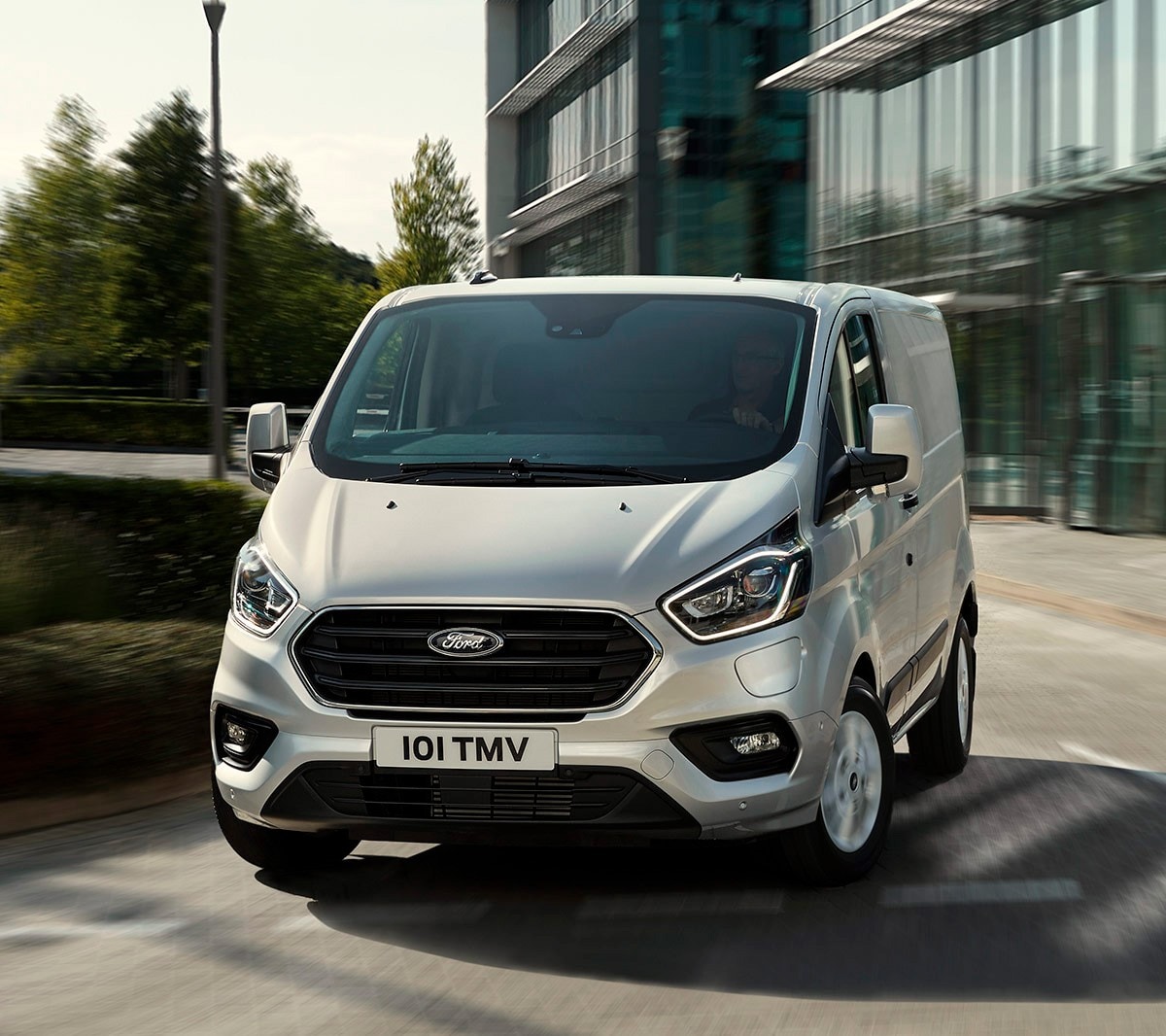 Silver Ford Transit Custom driving in front of glass building