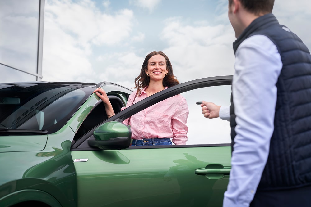 A woman in a pink shirt smiling beside a green car, with a Ford dealer handing her car keys.