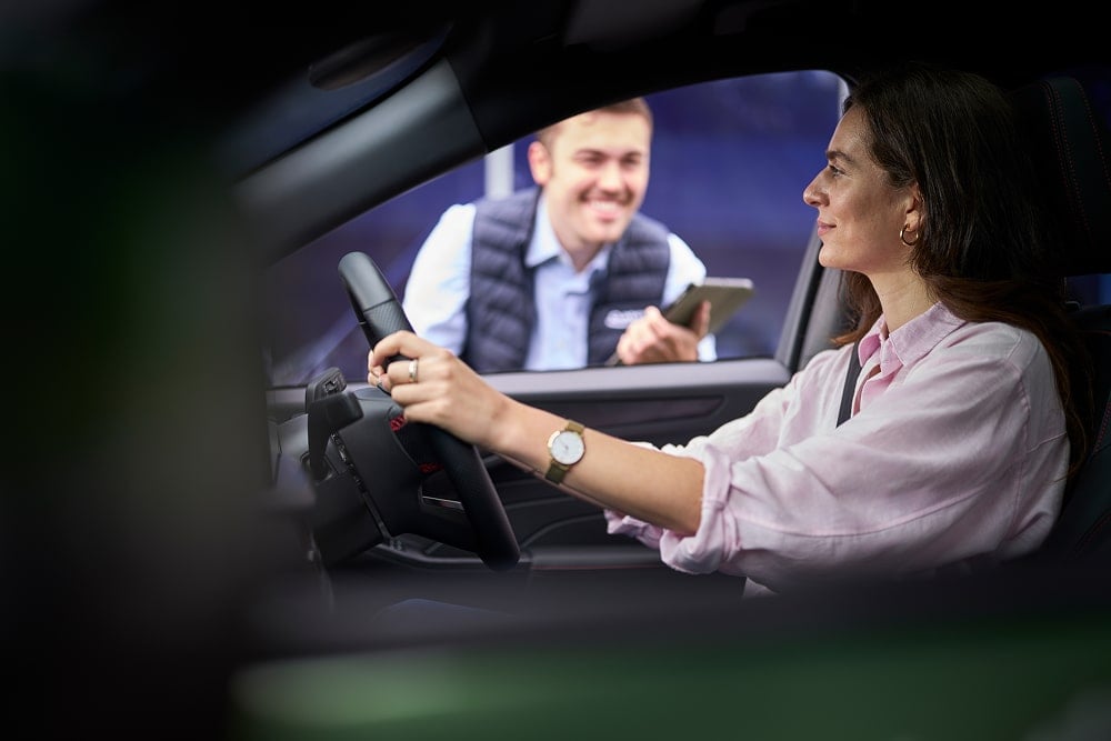 A woman in a parked green car smiling at a Ford dealer outside the window.