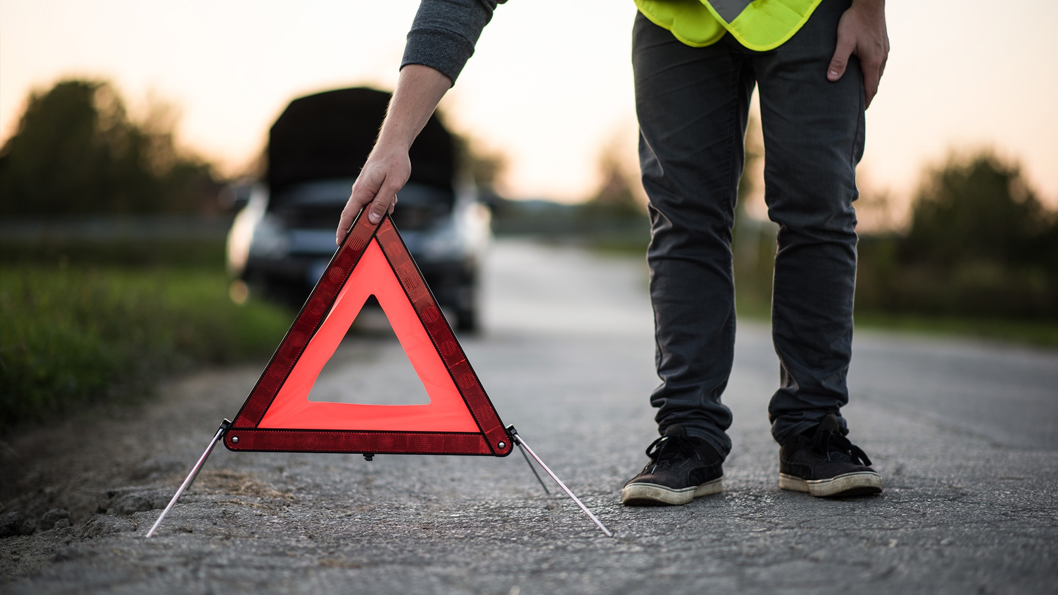 A person places a red warning triangle by a car that has broken down at the side of a road.