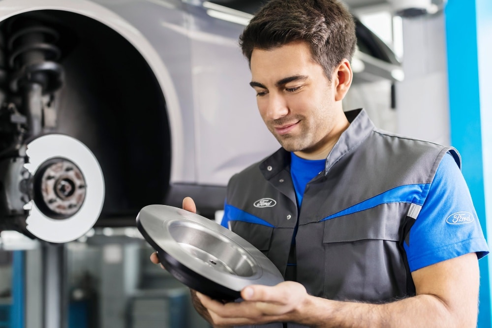 A technician in a Ford uniform inspects a new brake disc, with a car's brake assembly visible in the background