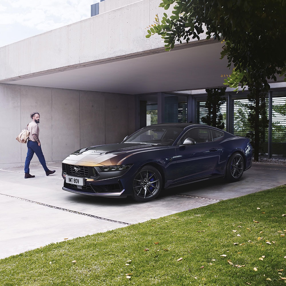 1000x1000 image of a navy blue Ford Mustang parked on a driveway as a person walks by checking it out.