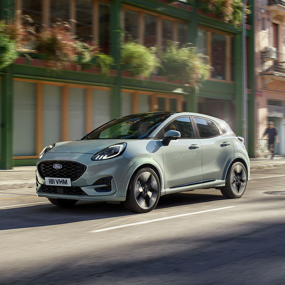 1000x1000 image of a grey Ford Puma driving on a main road.