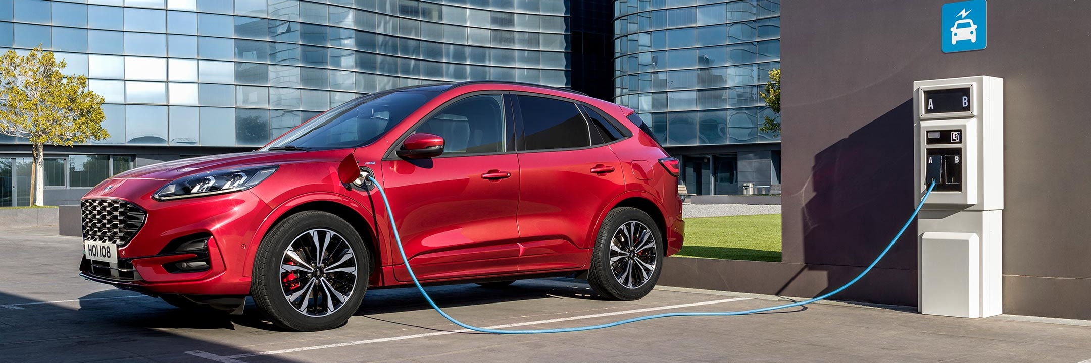Red Kuga Hybrid Electric Vehicle charging at a public charging point in front of glass buildings