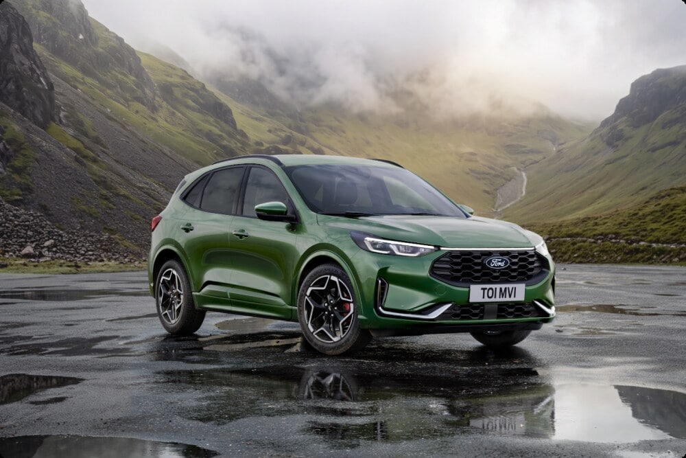 A green Ford Kuga parked on a paved promenade by the sea, with two people walking nearby.