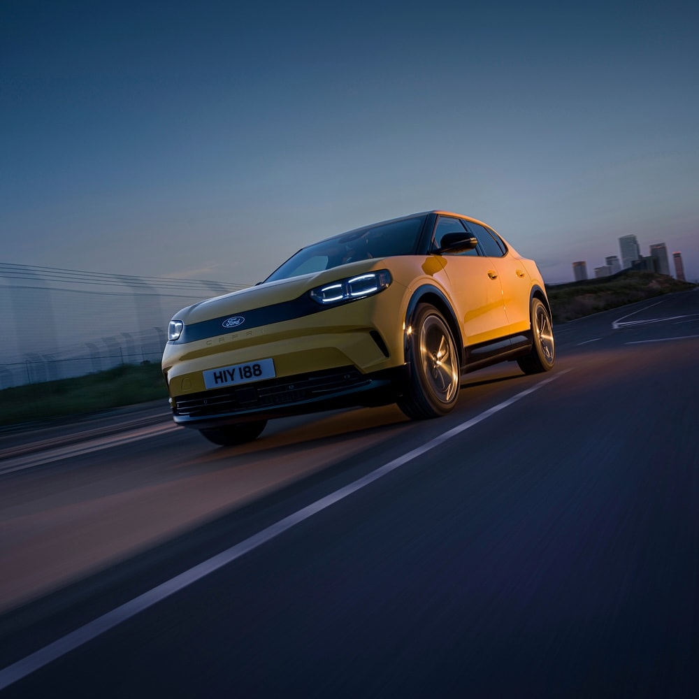 Yellow Ford Capri: Front 3/4 view of a yellow Ford Capri driving on a road at night with city lights.