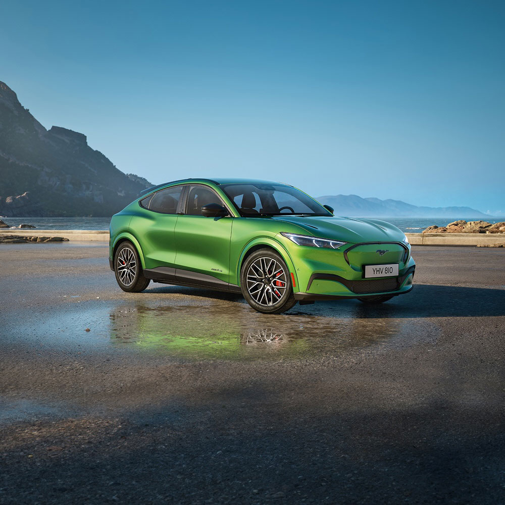 Green Ford Mustang Mach-E: Front 3/4 view of a green Ford Mustang Mach-E on a wet surface with mountains in the background.