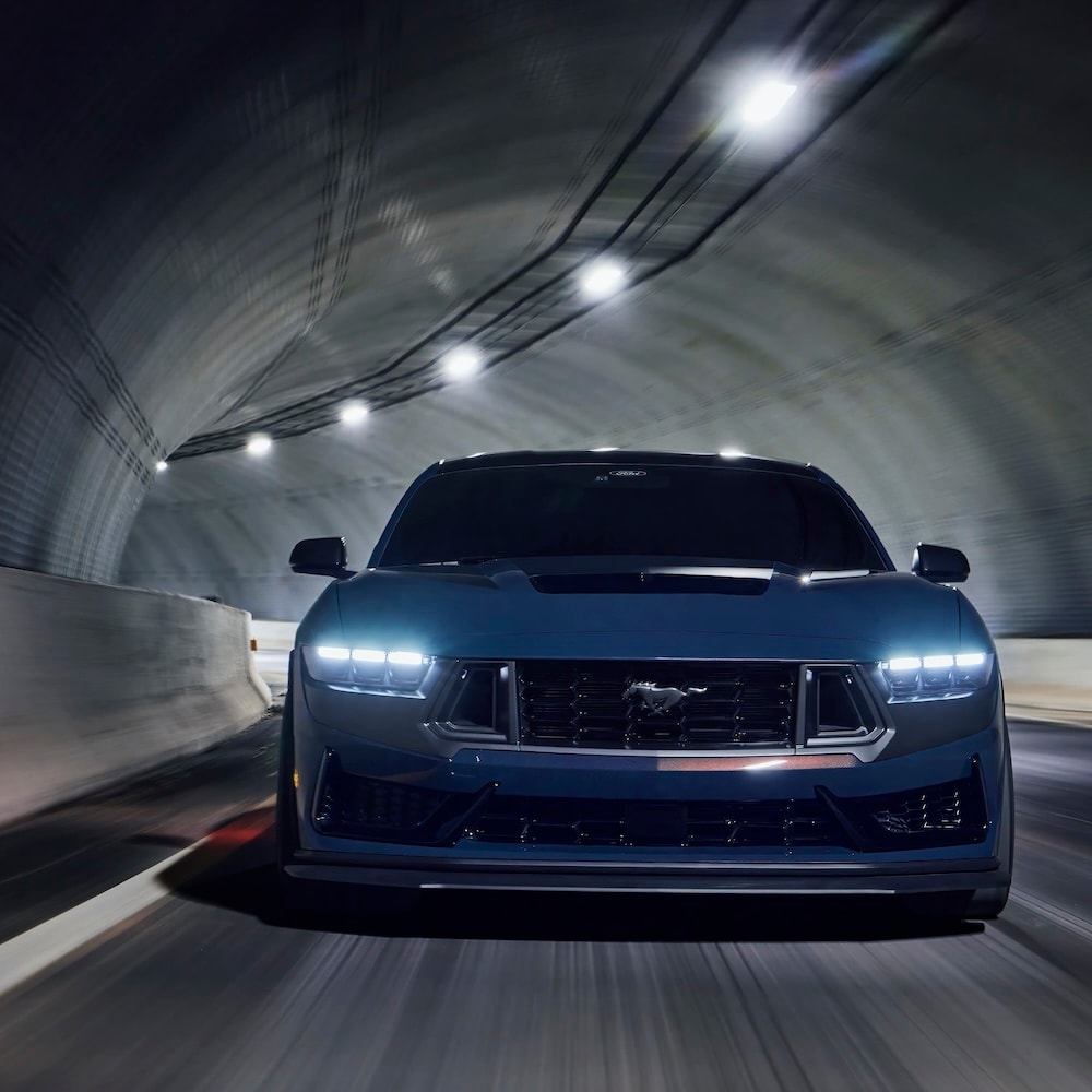 Front view of a dark blue Ford Mustang driving through a tunnel with its headlights on.