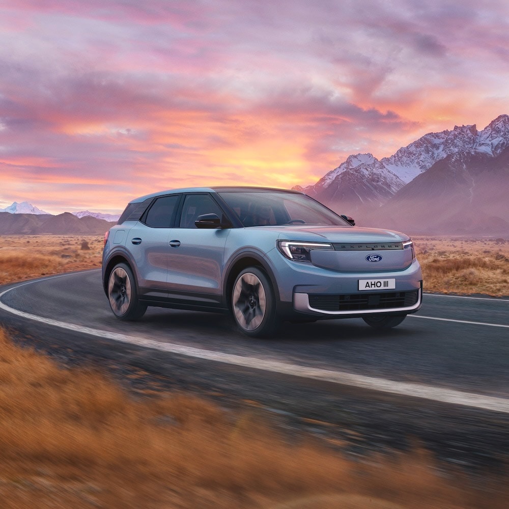 Light Blue Ford Explorer EV: Front 3/4 view of a light blue Ford Explorer EV driving on a mountain road at sunset.