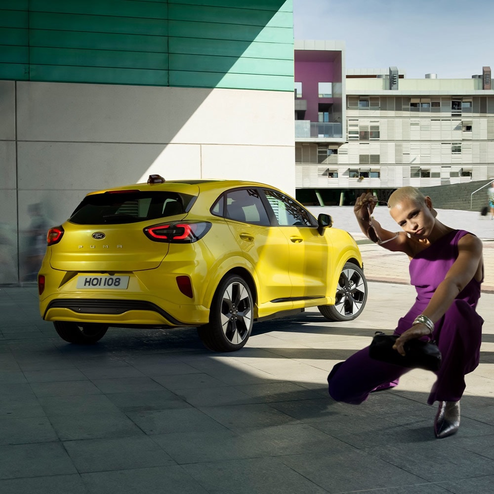A yellow Ford Puma Gen-E parked in front of a building, with a person crouching in the foreground.