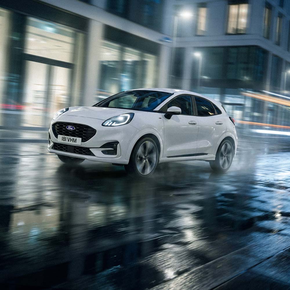 A white Ford Puma driving on a wet city street at night, seen from the front three-quarter angle.