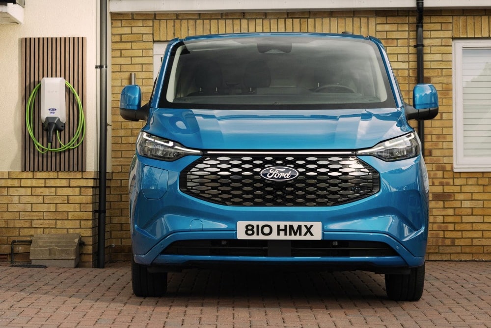 Front view of a blue Ford E-Transit Custom parked outside a brick house with an EV charger on the wall.