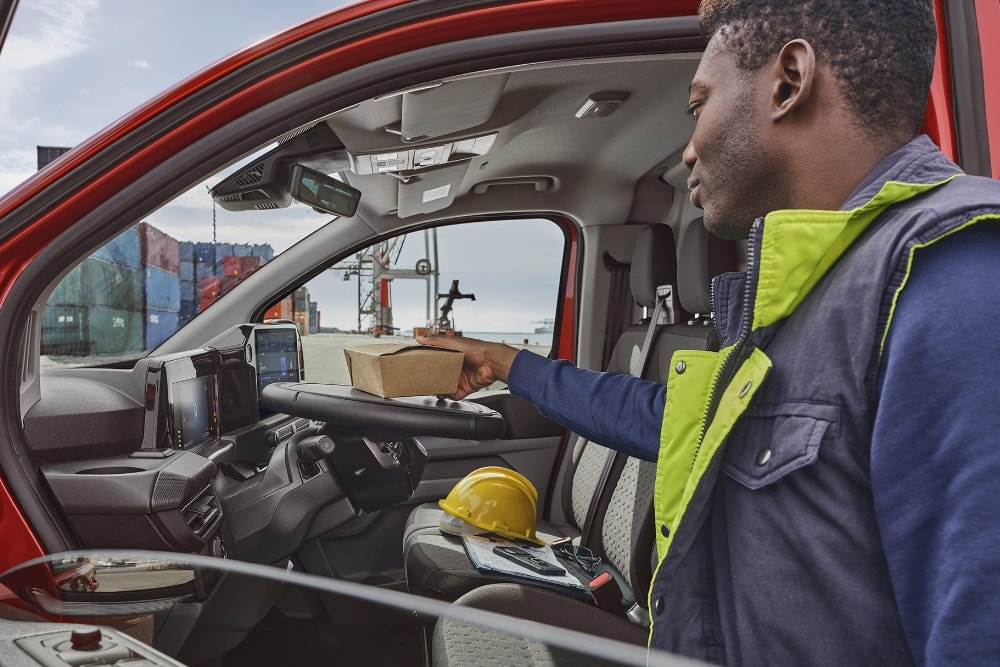 A person hands a package into a red Ford Transit Custom van, with shipping containers visible in the background.