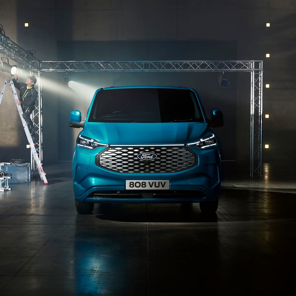 Front view of a Ford E-Transit Custom in a studio, with a person on a ladder in the background.