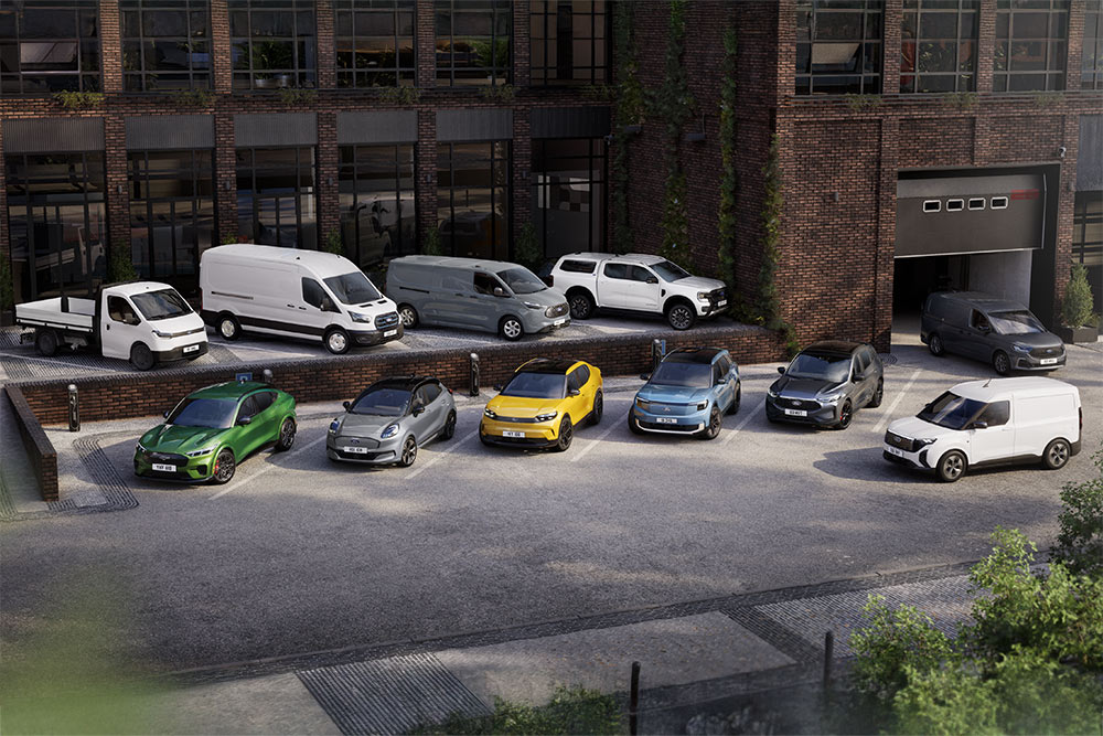 A range of Ford passenger cars and commercial vehicles parked outside a brick building.
