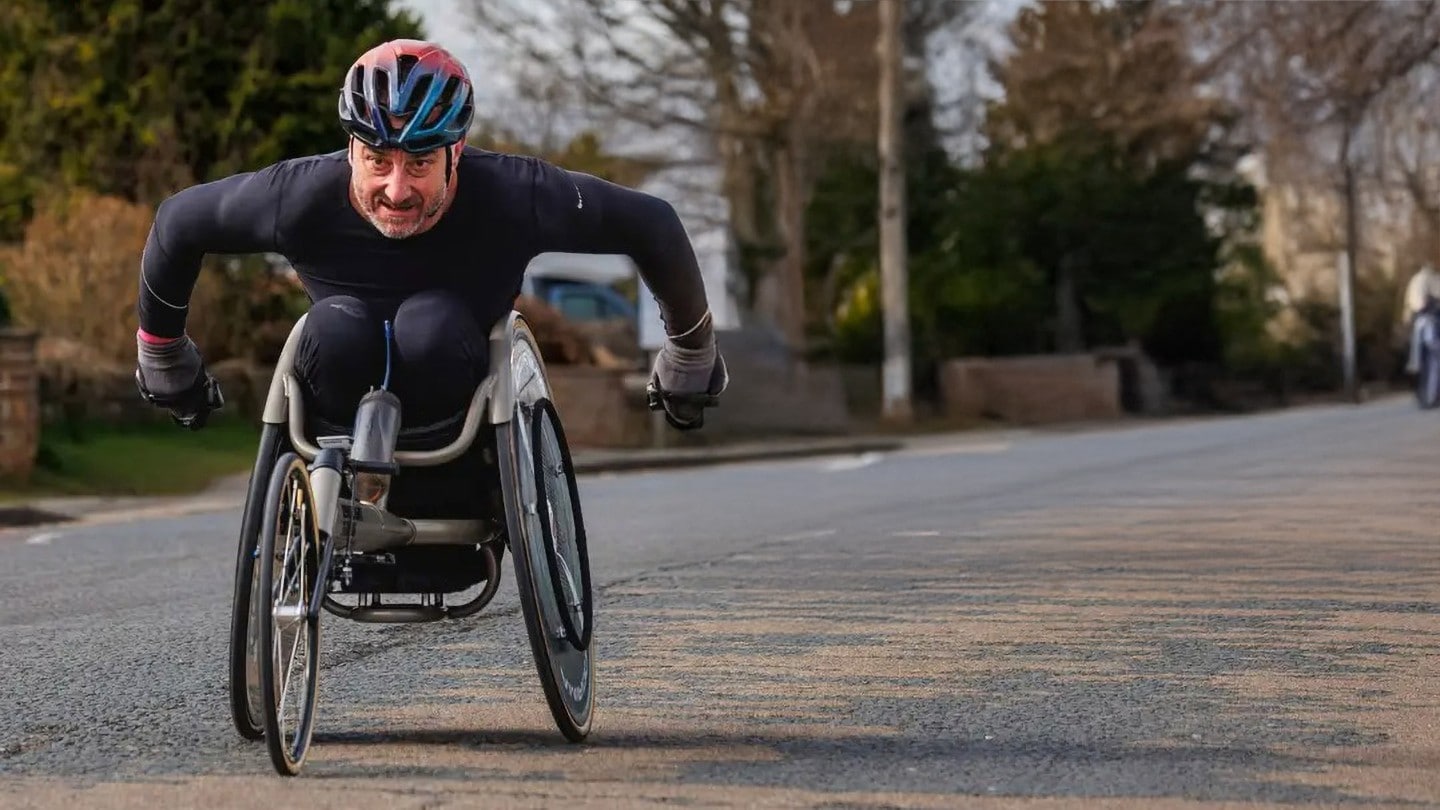 Jason Richards in a racing wheelchair wearing a helmet, propelling themselves along a road