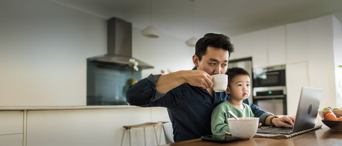 A person holding a child, working on a laptop and drinking from a mug at a table.