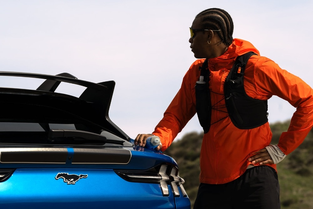 A person in an orange jacket stands beside the rear spoiler and badge of a blue Ford Mustang Mach-E.