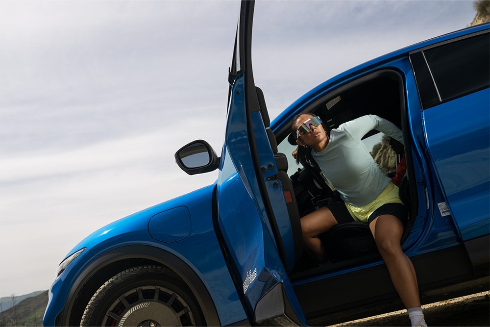 A person steps out of an open door of a blue Ford Mustang Mach-E parked outdoors.