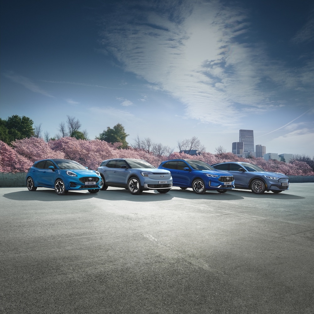 Four Ford vehicles: a blue Puma, light blue Explorer, dark blue Kuga, and grey Mustang Mach-E, parked against cherry blossom trees and a city skyline.