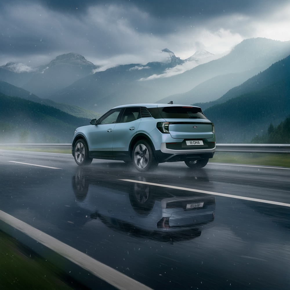 Rear 3/4 view of a light blue Ford Capri driving on a wet road with mountains in the background.