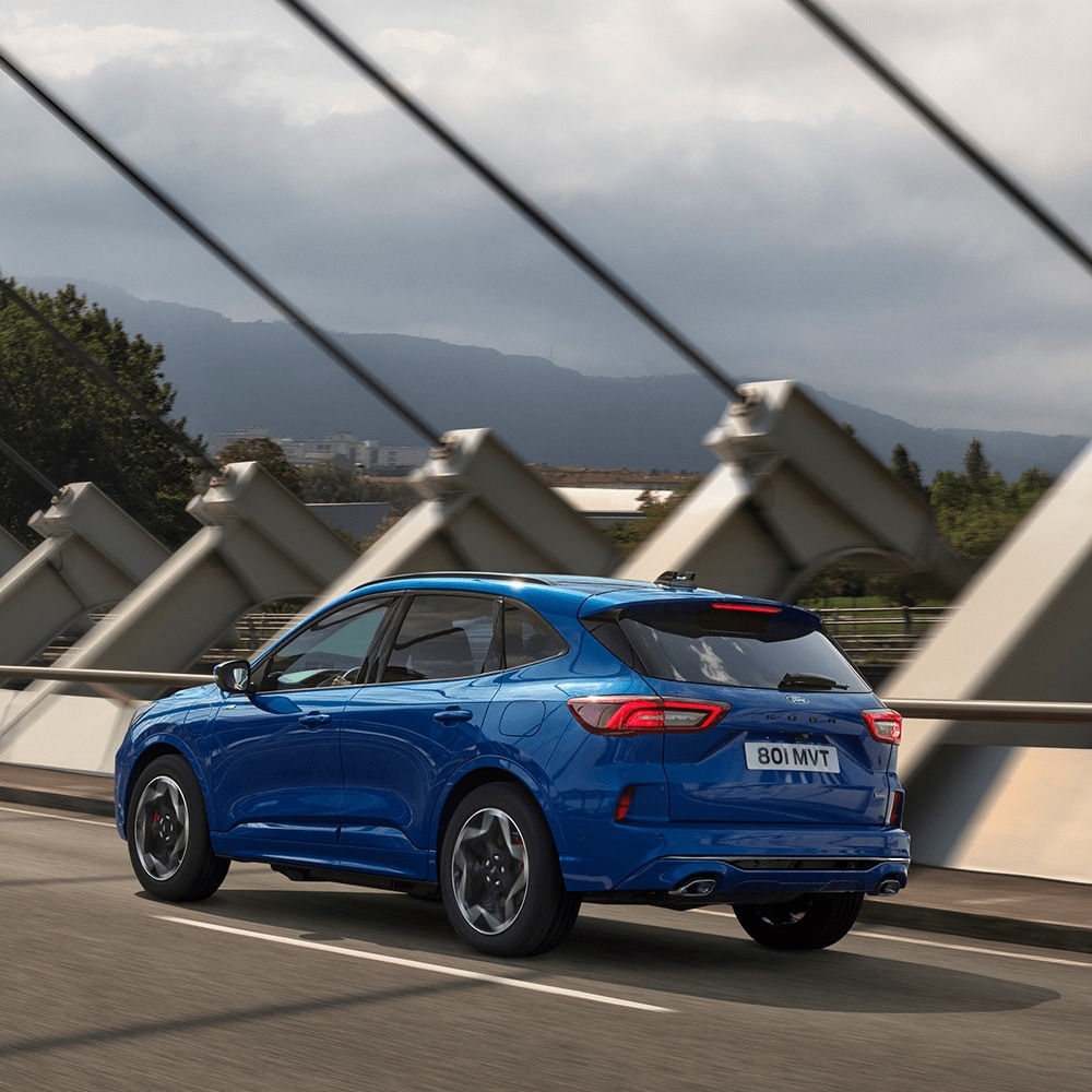 A blue Ford Kuga driving on a bridge, with mountains in the background.