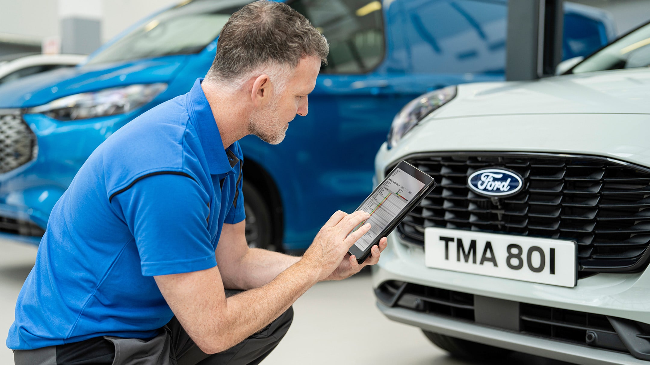 Technician kneeling, using a tablet to inspect the front of a white Ford car.