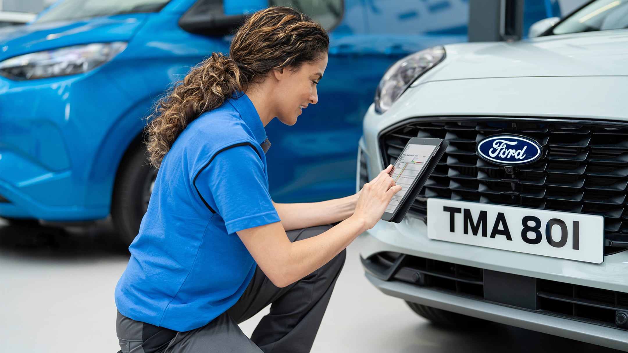 Technician kneeling, using a tablet to inspect the front of a white Ford car.