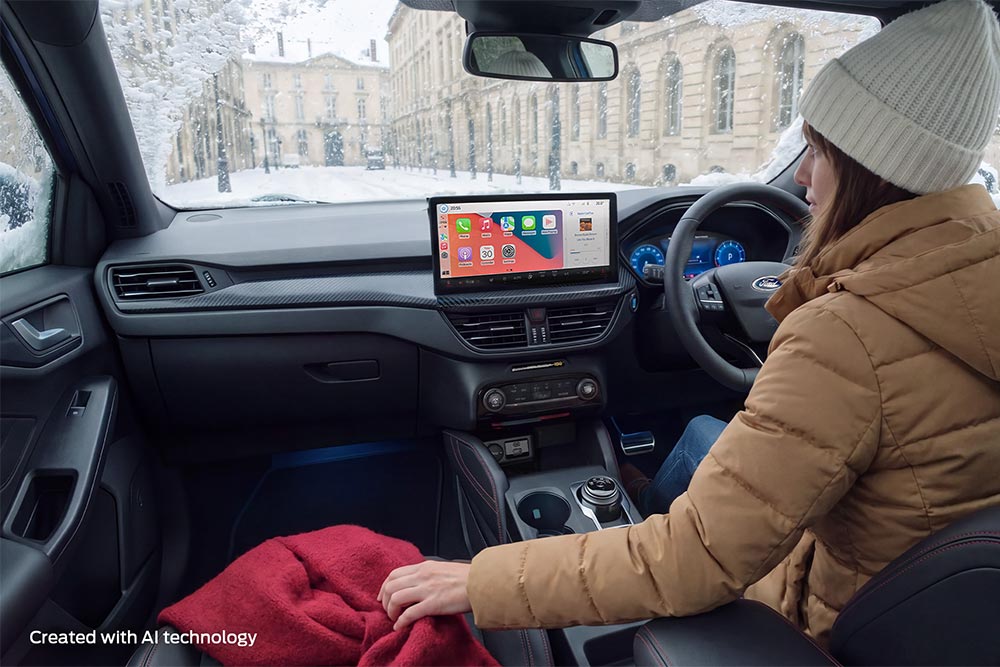 A woman wearing a winter coat and beanie sits in a Ford car on a snowy day, holding a red blanket.