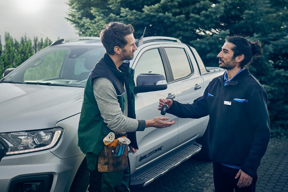 Technician and customer exchange keys in front of a silver Ford Ranger pickup truck parked outdoors.