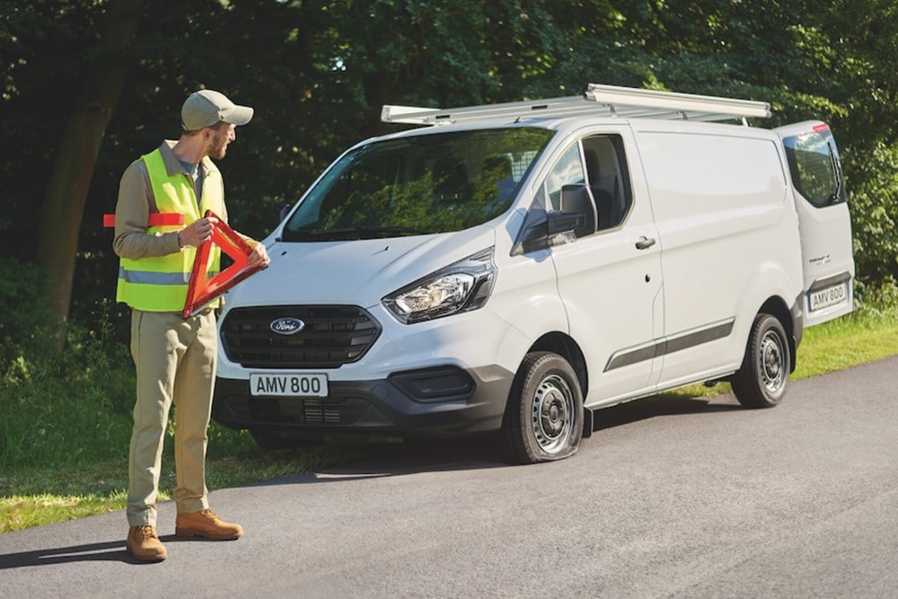A person in a high-visibility vest places a warning triangle beside a white Ford van on a country road.